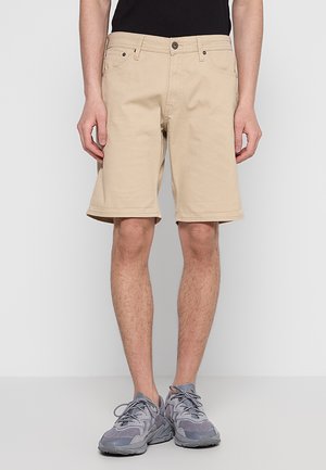 Mann trägt beige knielange Shorts, schwarzes Hemd und graue Sportschuhe, steht vor einem einfarbigen hellgrauen Hintergrund.