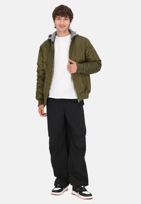 Oliven grøn bomberjakke med grå hætte, hvid t-shirt, sorte bukser og sorte sneakers med røde accenter. Modellen står afslappet med et smil.