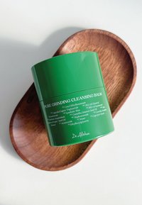 Zielony cylindryczny pojemnik z napisem "Pure Grinding Cleansing Balm" w białym kolorze, umieszczony na brązowej, owalnej drewnianej tacy.