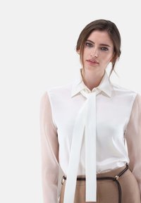 Camicia bianca con maniche lunghe trasparenti e un dettaglio di cravatta al collo. Tessuto liscio con un design aderente, abbinata a una gonna marrone a vita alta.