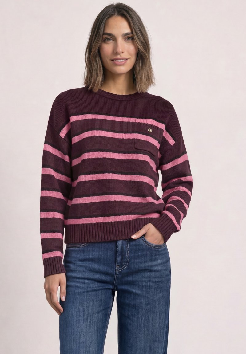 Femme portant un pull rayé bordeaux et rose et un jean bleu, debout avec une main dans la poche devant un fond uni.