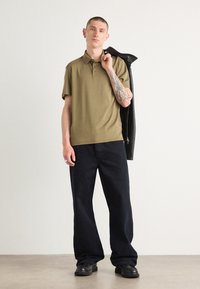 Polo shirt vert olive à manches courtes et trois boutons, associé à un pantalon large bleu foncé et des chaussures noires. Veste en cuir drapée sur l'épaule.