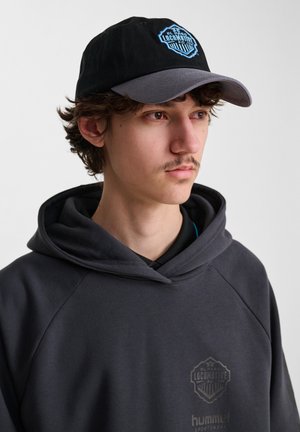 Sort baseball cap med grå skygge, der har et blåt og sort broderet logo. Grå sweatshirt med et diskret logo på brystet.