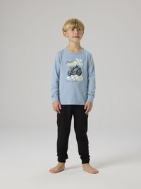 Νέος αγόρι όρθιος ξυπόλυτος, φορώντας ανοιχτό μπλε μπλουζάκι με μακρύ μανίκι και γραφικό μοτοσικλέτας και μαύρο παντελόνι, μπροστά σε γκρι φόντο.