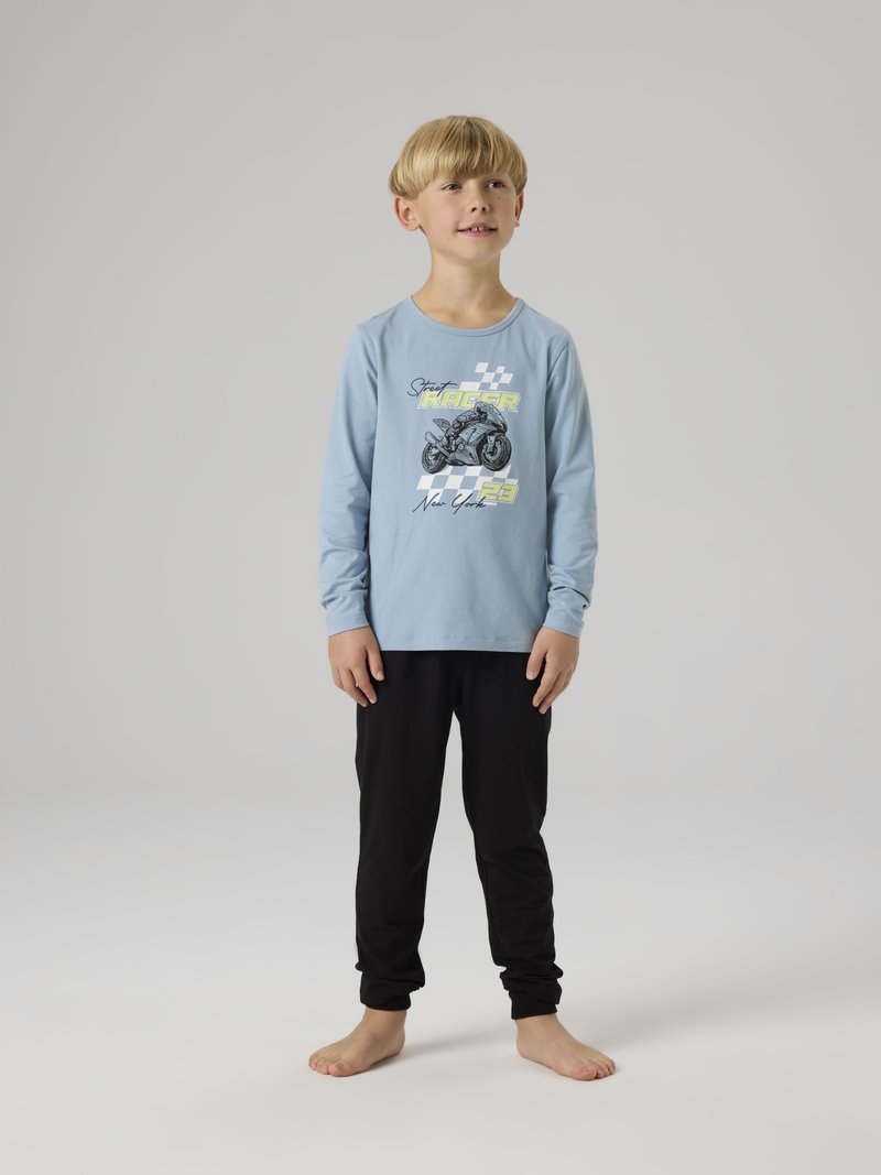 Νέος αγόρι όρθιος ξυπόλυτος, φορώντας ανοιχτό μπλε μπλουζάκι με μακρύ μανίκι και γραφικό μοτοσικλέτας και μαύρο παντελόνι, μπροστά σε γκρι φόντο.