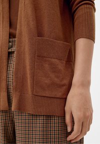 Hnědý svetr na knoflíky z měkkého materiálu s žebrovanými manžetami a přední kapsou, ve spojení s károvanými hnědými a béžovými kalhotami.
