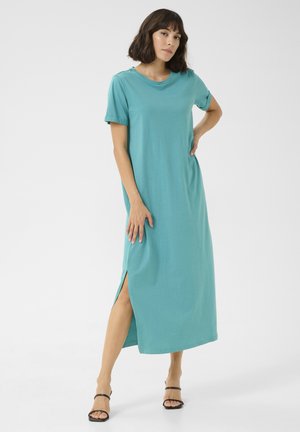 Tealfarbendes Baumwoll-Maxikleid mit kurzen Ärmeln, Rundhalsausschnitt und seitlichen Schlitzen. Das Model posiert mit einer Hand an der Hüfte und trägt schwarze Riemchensandalen.
