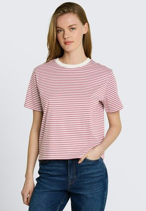 Jeune femme aux cheveux châtain clair raides portant un t-shirt à manches courtes rose et blanc à rayures et un jean bleu, main dans la poche.