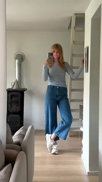 Hellgraues Langarmoberteil kombiniert mit weit geschnittenen blauen Jeans und weißen Sneakers. Die Umgebung umfasst einen Spiegel und einen schwarzen Holzofen.