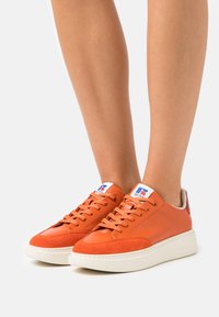 Orange lädersneakers med en vit gummisula, med en mocka tåhuvud och en logotyp på tungan. Snörningarna är orange; hälen har en röd accent.