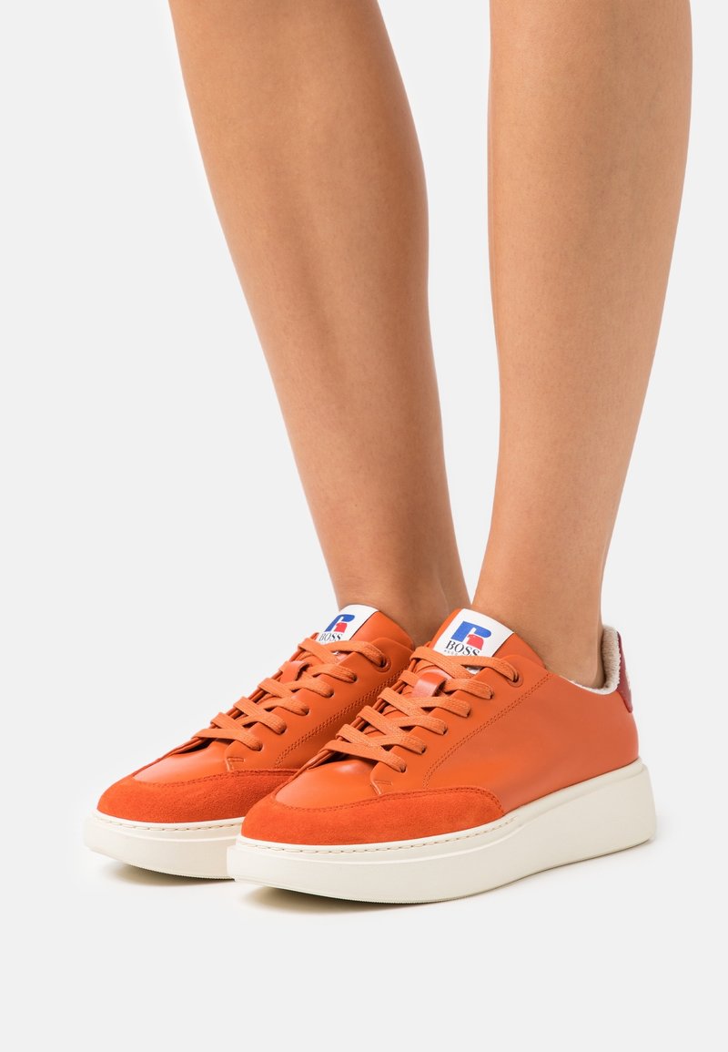 Orange lädersneakers med en vit gummisula, med en mocka tåhuvud och en logotyp på tungan. Snörningarna är orange; hälen har en röd accent.