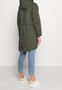 Parka de color verde oliva con capucha, cintura ajustada, abertura trasera y detalles en neón contrastantes, combinada con vaqueros de mezclilla azul claro y zapatillas blancas.