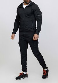 Haut de survêtement zippé noir avec capuche à cordon de serrage et poches zippées. Associé à un pantalon cargo noir avec des poches latérales. Baskets noires avec des accents orange.