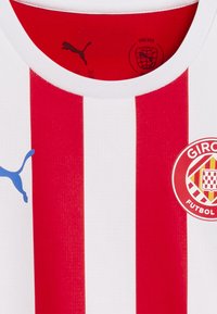Weißes Trikot mit roten vertikalen Streifen. Verfügt über ein blaues Puma-Logo und ein rotes kreisförmiges Girona FC-Emblem mit Schachbrettmuster. Rippkragen.
