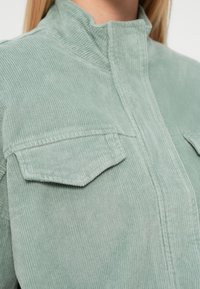 Chemise en velours côtelé vert avec col montant, deux poches poitrine et texture côtelée. Tissu doux avec finitions de couture soignées.