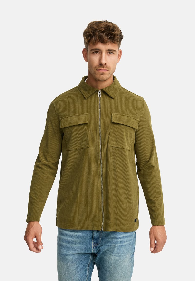 Olivengrøn kordjakke med lynlås foran, flad krave og to brystlommer; båret over blå denimjeans. Glat tekstur.