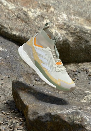 Pantof de drumeție Adidas Terrex gri deschis și galben, în aer deasupra unui teren stâncos cu particule de pământ împrăștiate în jur.