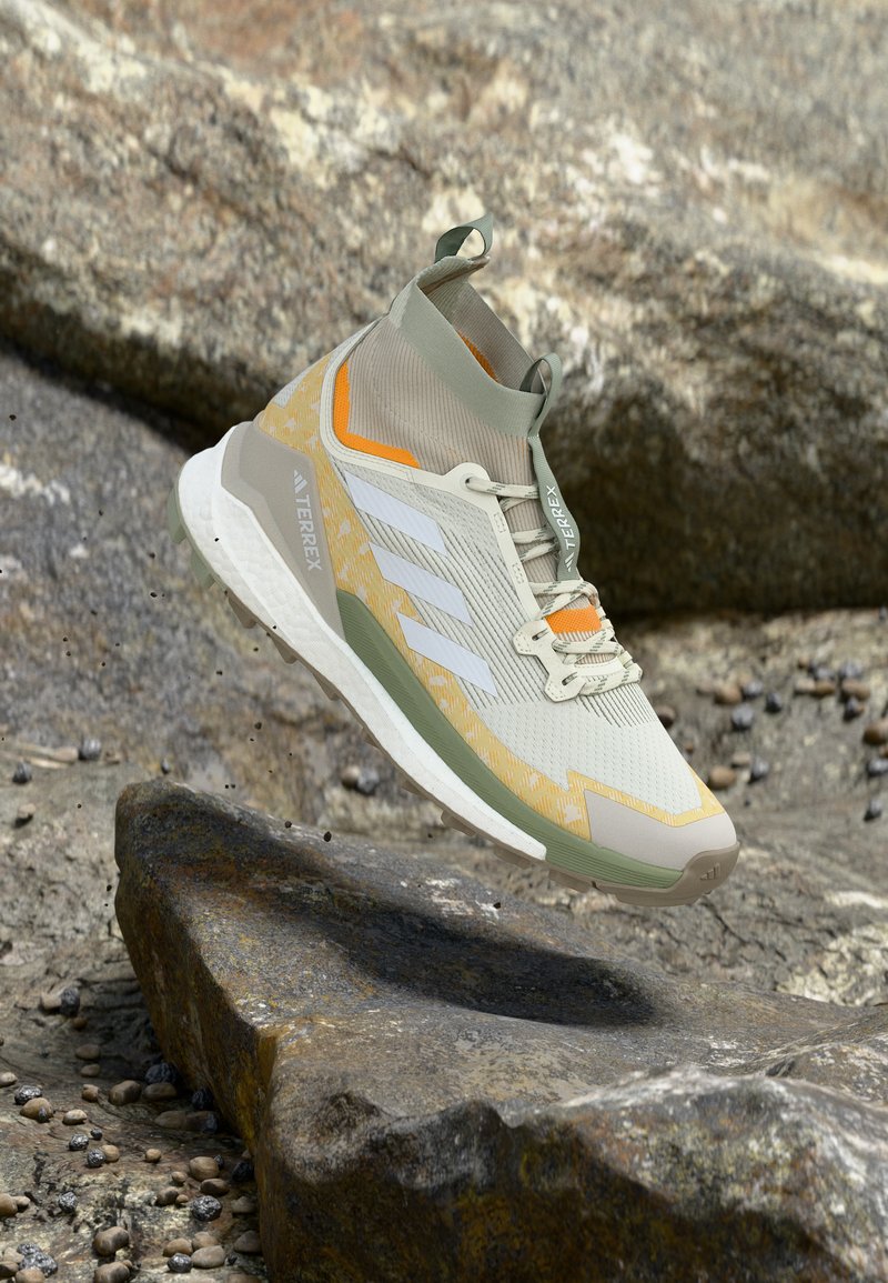 Lichtgrijze en gele Adidas Terrex wandelschoen in de lucht boven rotsachtig terrein met rondvliegende vuildeeltjes.