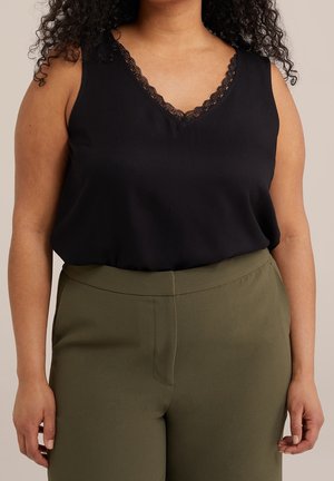 Femme portant un haut noir sans manches avec bordure en dentelle et un pantalon taille haute vert olive, sur un fond neutre.