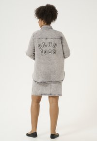 Grå denimjakke med "CLUB 1993" broderi på ryggen, parret med en matchende denimnederdel. Glat tekstur, afslappet pasform og rå kant.