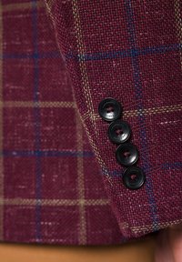Bordowy materiał tweedowy w kratę z niebieskimi i beżowymi liniami, ozdobiony czterema czarnymi guzikami na mankiecie.