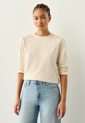 Camicia beige a maniche lunghe in tessuto morbido con scollo rotondo, abbinata a jeans azzurri a vita alta, che mostrano una vestibilità casual.