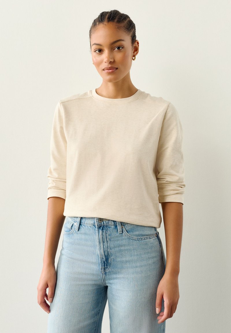 Beigenvärinen pitkähihainen paita, valmistettu pehmeästä kankaasta, pyöreällä pääntiellä, yhdistetty vaaleansinisiin korkeavyötäröisiin farkkuihin, jotka esittelevät rentoa istuvuutta.