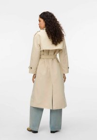 Beige trenchcoat med dubbla knappar, bälte i midjan, ryggslits och axelremmar, tillsammans med löst sittande ljusblå jeans.