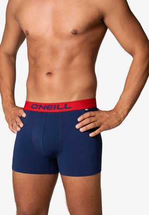 Mannelijke torso met marineblauwe boxershorts met een rode tailleband met het merk "O'NEILL", handen op de heupen tegen een witte achtergrond.