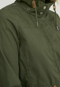 Olivově zelená bunda s vysokým límcem, předním zapínáním na cvoky, nastavitelnými šňůrkami a kontrastními hnědými detaily na kovových částech.