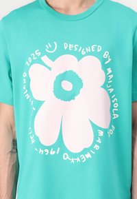 Turquoise cotton t-shirt featuring a large white flower graphic with text around it. Soft texture, crew neck design, and relaxed fit.