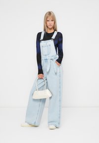 Lichtblauwe denim salopettes met wijde broekspijpen, verstelbare banden en een voorkantzak, gecombineerd met een aansluitend zwart topje en een kleine witte handtas.