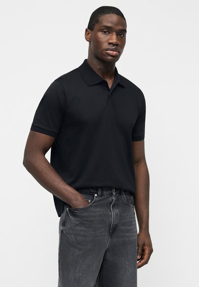 Άνδρας που φοράει μαύρο πόλο μπλουζάκι με κοντά μανίκια και γιακά, μαζεμένο ελαφρώς μέσα σε ξεθωριασμένο μαύρο τζιν, στέκεται με το ένα χέρι στην τσέπη.