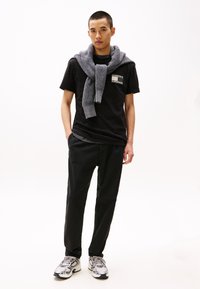 Camiseta negra con un bolsillo de parche, suéter gris de punto caído sobre los hombros, pantalones negros y zapatillas deportivas gris/plata. Look sencillo y moderno.