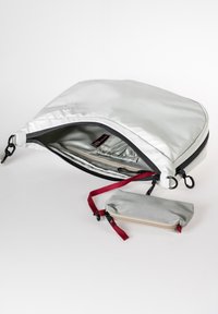 Weiße Stofftasche mit glatter Textur, ausgestattet mit einem schwarzen Reißverschluss und einem roten Tragegurt, zusammen mit einem kleinen grauen Beutel mit Reißverschluss.