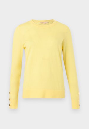 Hosszú ujjú, halvány sárga pulóver kerek nyakkivágással és három barna gombbal minden mandzsettán, világos szürke háttéren.