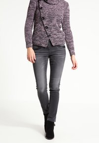 Grijze denim skinny jeans gecombineerd met een gestructureerde paarse gebreide trui met grote zwarte knopen en een kraaghals, gedragen met zwarte hoge hakken.