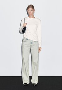 Bílá košile s dlouhými rukávy v kombinaci s širokými džíny s bledým praním. Modelka drží malou černou kabelku. Minimalistický design bez vzorů.