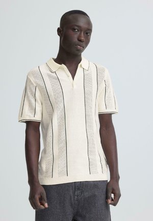 Giovane uomo nero che indossa una polo lavorata a maglia color crema a maniche corte con righe verticali nere e jeans grigio scuro, in piedi davanti a uno sfondo semplice.