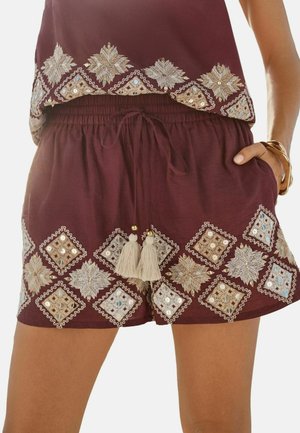 Frau, die bordeauxrote Shorts und ein Top mit beigen und weißen bestickten geometrischen Mustern sowie Quasten-Kordelzügen an den Shorts trägt.