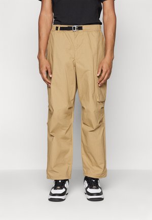 Beige cargo pants with a relaxed fit, adjustable black belt, textured fabric, and side pockets. Worn with black and white sneakers.