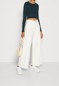 Weiße weite Jeans, dunkelgrünes, geripptes Cropped Top und weiße Sneakers. Als Accessoire dient eine kleine beigefarbene Handtasche mit goldenen Akzenten.