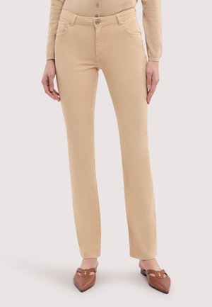 Mujer con pantalones beige de tiro recto, camiseta beige de manga larga y zapatos planos marrones de punta puntiaguda con detalle de hebilla dorada.