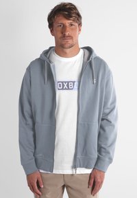 Veste à capuche zippée grise avec un cordon de serrage, deux poches avant, des poignets côtelés, et un T-shirt blanc portant un design graphique bleu et noir en dessous.