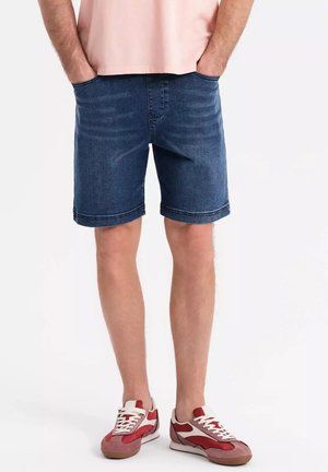 Shorts en denim bleu foncé, avec une coupe classique, cinq poches et un ourlet propre. Portés avec des baskets rouges et blanches et une chemise rose clair.