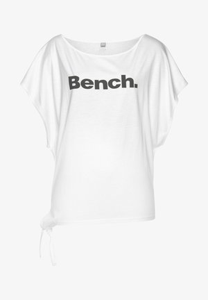 T-shirt oversized blanc avec un large col et des manches courtes, présente un logo noir "Bench." très visible. Comprend un détail de nouage sur le côté.