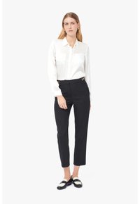 Camisa blanca de manga larga con dos bolsillos en el pecho, combinada con pantalones negros de cintura alta con detalles dorados y con dobladillos cortos. Zapatos en blanco y negro.