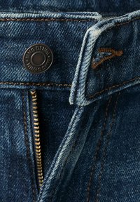 Jean en denim avec fermeture éclair partiellement ouverte, bouton en métal marqué "Denim" et détails de surpiqûres orange contrastantes.