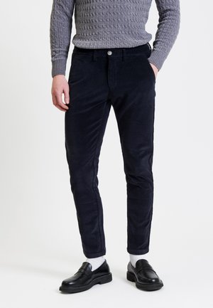 Granatowe spodnie z korduroju o slim fit, z przednimi kieszeniami, paskami na pasek i zapięciem na guzik. W zestawieniu z czarnymi loafersami i szarym swetrem.