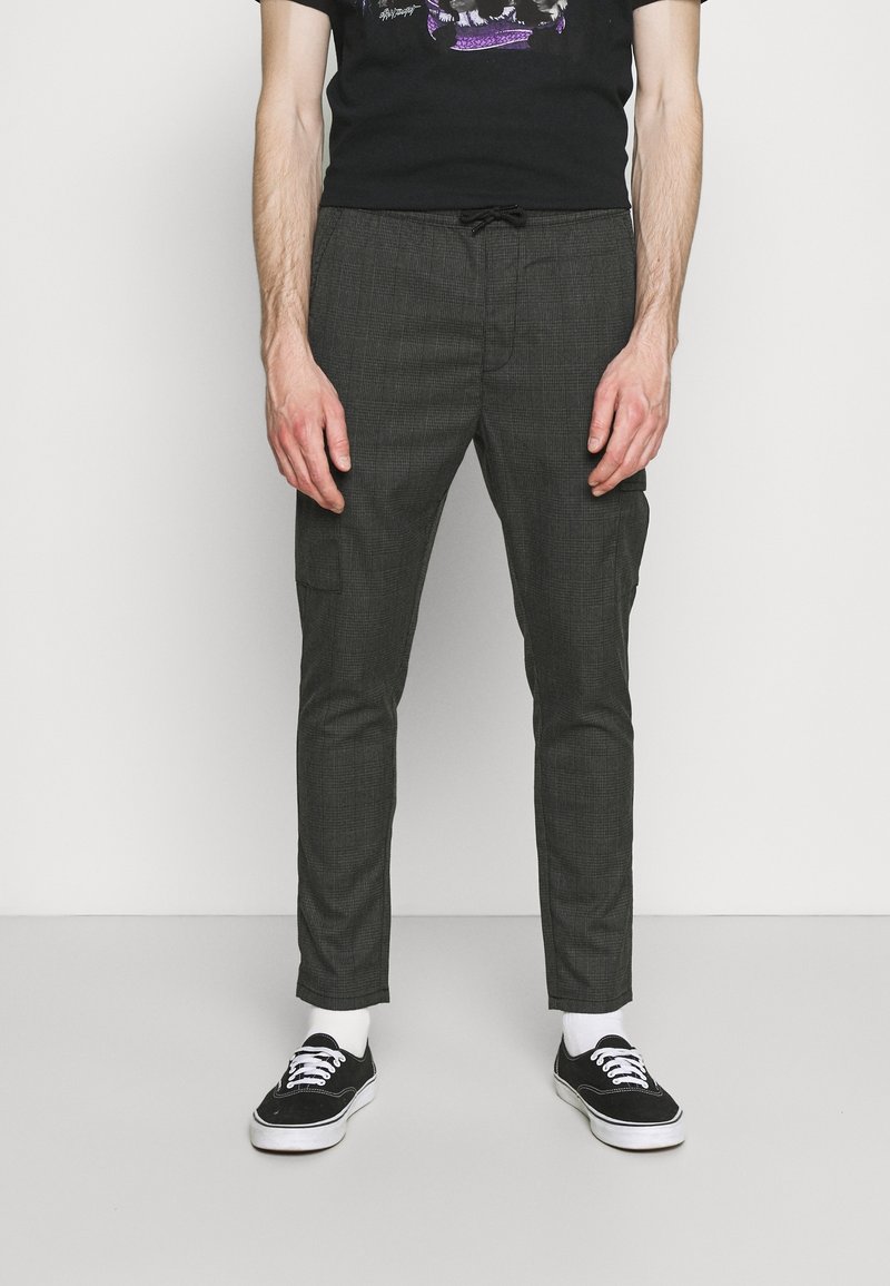 Homme portant un pantalon slim à carreaux gris, un t-shirt noir avec un motif graphique, des chaussettes blanches et des baskets noires, debout devant un fond blanc.